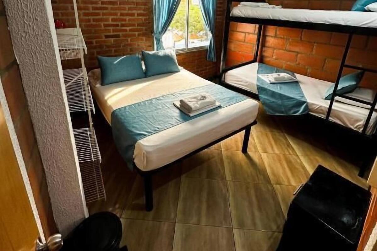 A compact room with a bunk bed, a single bed, a window with curtains, and a small shelf unit.