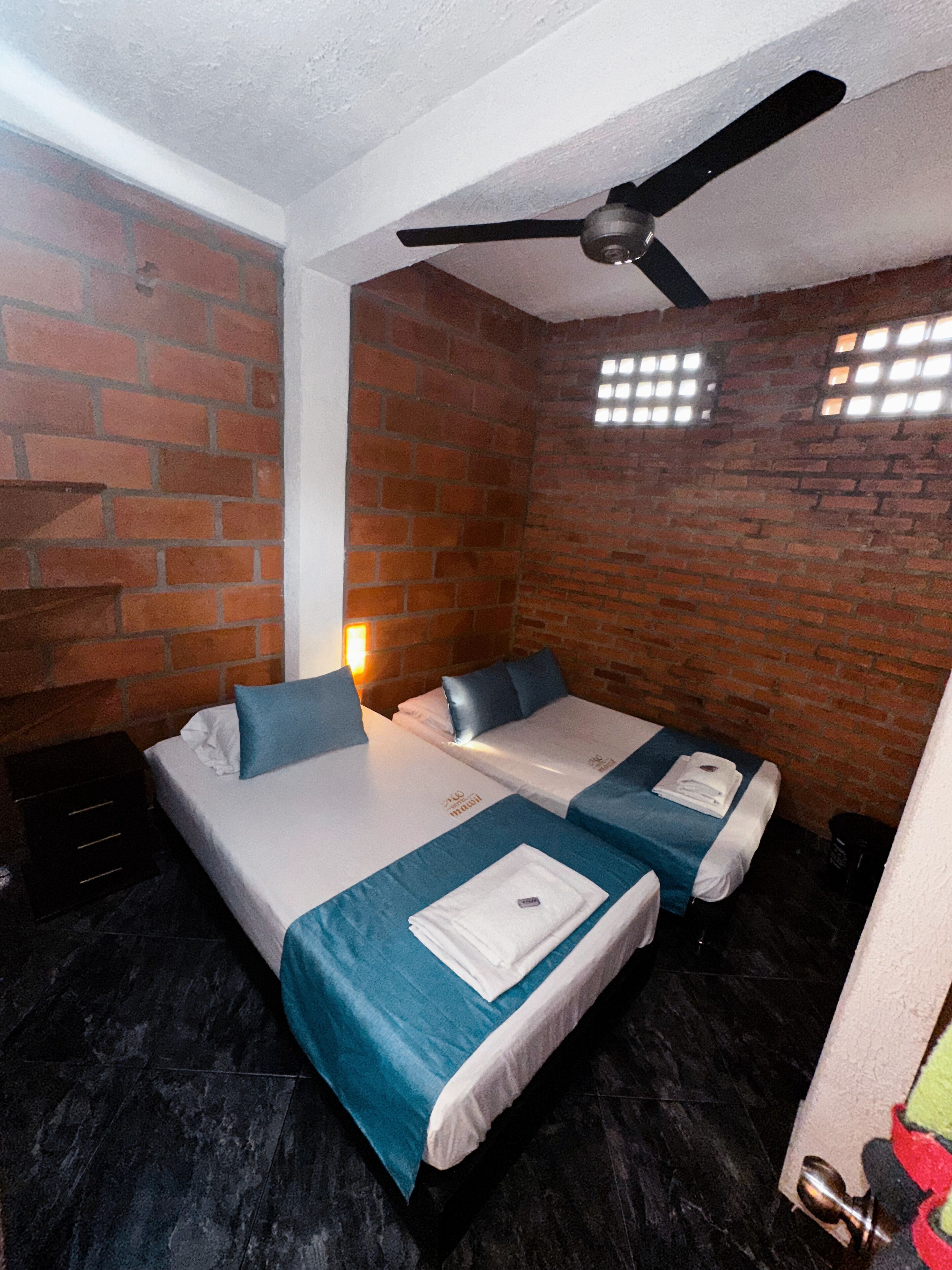 A room with two beds, a ceiling fan, brick walls, and a tiled floor.