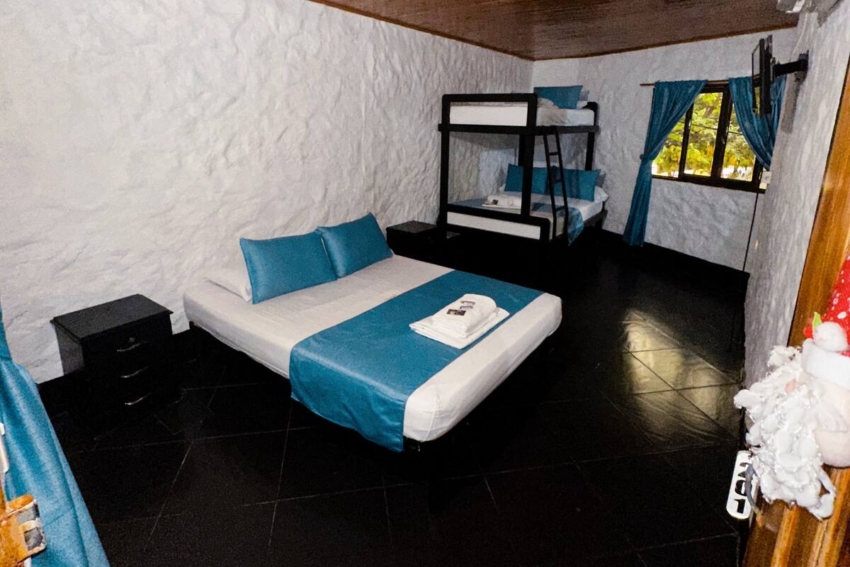 A compact room with a bunk bed, a single bed with blue bedding, a small bedside table, a window with curtains, and a wooden ceiling.