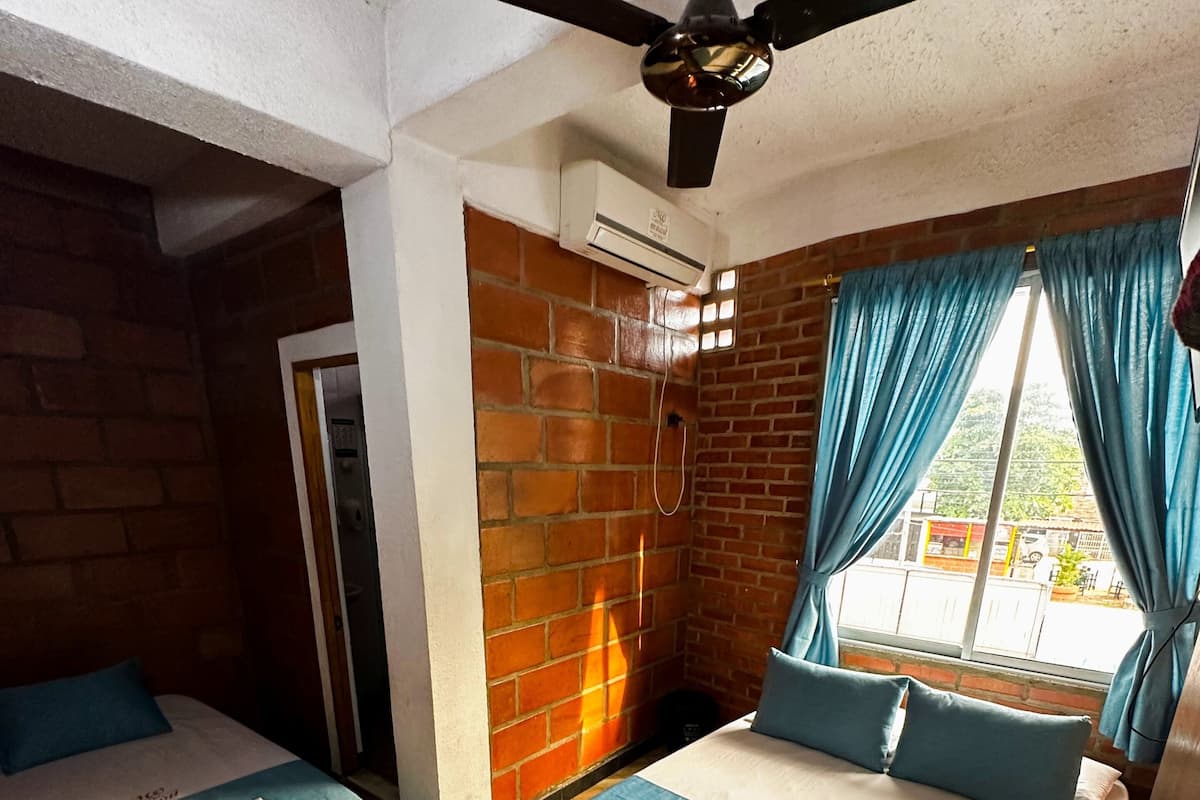 A hotel room with two beds, a ceiling fan, a brick wall, and a window with curtains.