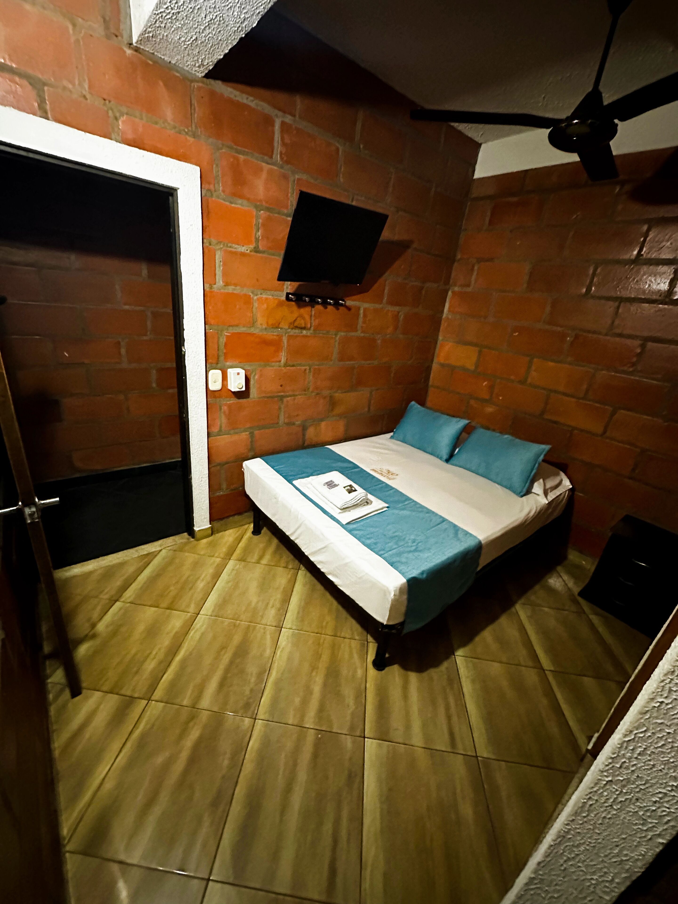 A small, cozy room with a brick wall, a bed with blue bedding, a flat-screen TV mounted on the wall, and a ceiling fan.