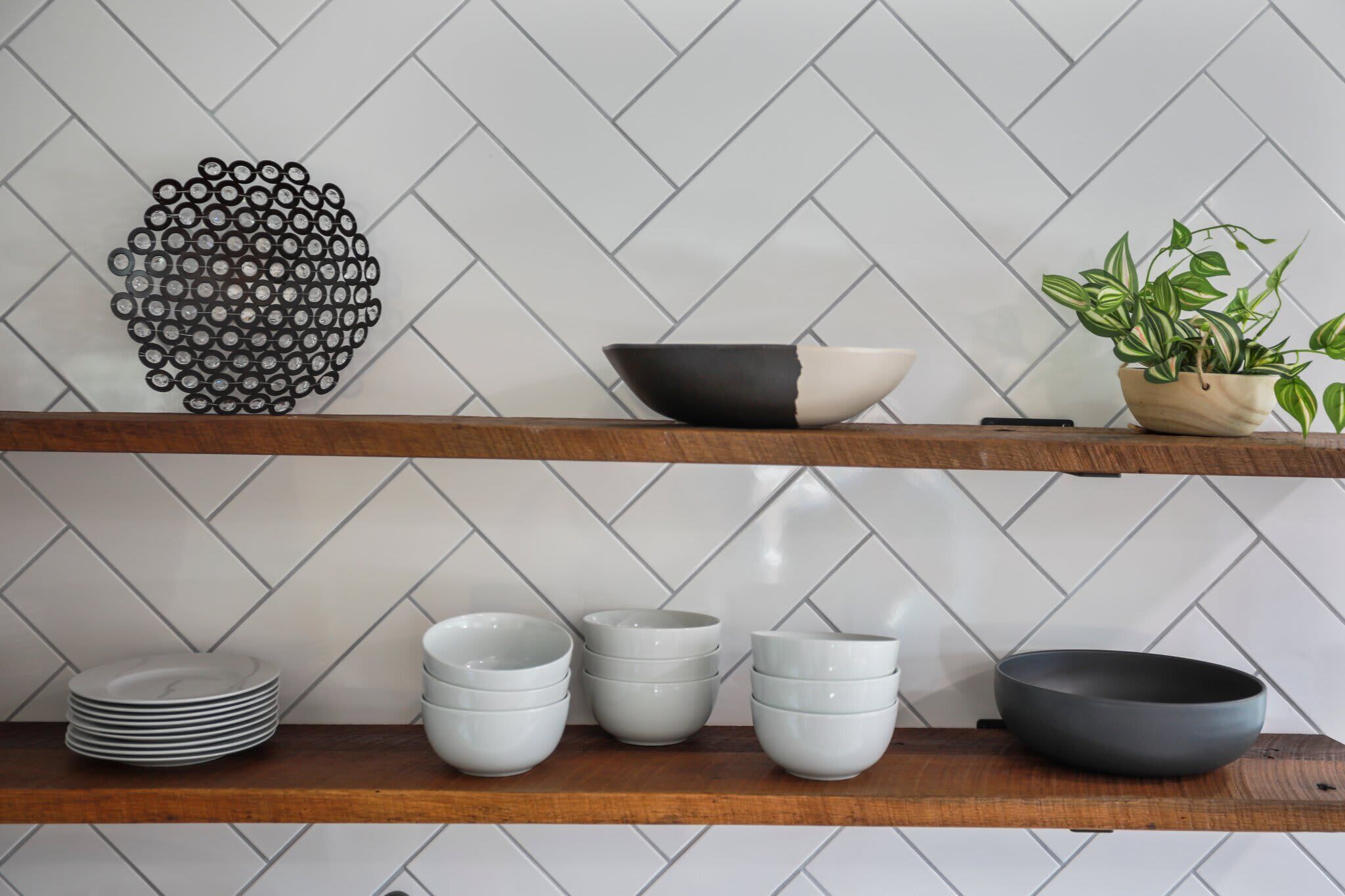 A kitchen with two wooden shelves, white herringbone-patterned backsplash, and various kitchenware including bowls and plates.