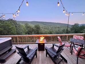 Una terraza con un hoyo para el fuego, mobiliario exterior e iluminación colgante, con un paisaje boscoso de fondo.