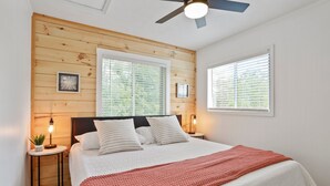 Une chambre avec un plafond en bois, un lit avec une literie blanche et une couverture rouge, deux tables de chevet avec des lampes et un ventilateur de plafond.