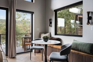 Una habitación moderna con una mesa redonda, dos sillas, un banco y un ventanal con vistas a un bosque.
