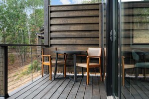 Una terraza de madera con una mesa, sillas, un ventanal y vistas a árboles.