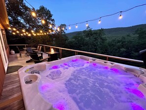 A hot tub with purple lighting on a wooden deck, surrounded by string lights and overlooking a forested area. - Starstone Chalet - Hot Tub! Mountain Views! (Broadway)