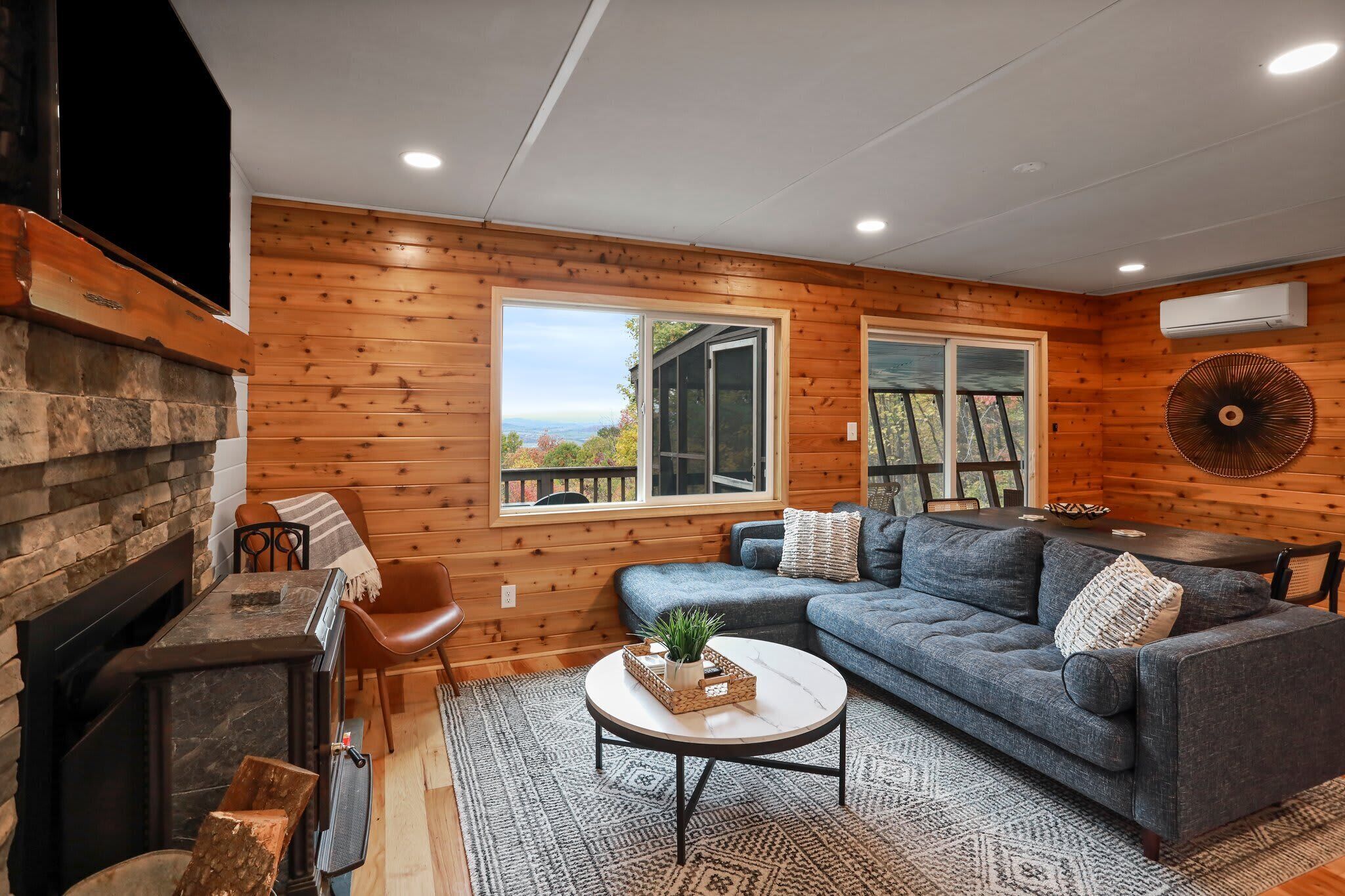 A cozy living room with a fireplace, a sectional sofa, a coffee table, and large windows offering a view of the outdoors.
