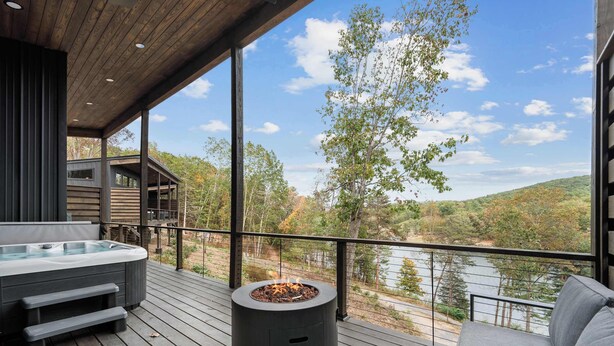 Una terraza de madera con hidromasaje, un pozo de fuego y un sofá con vistas a un lago y un bosque.