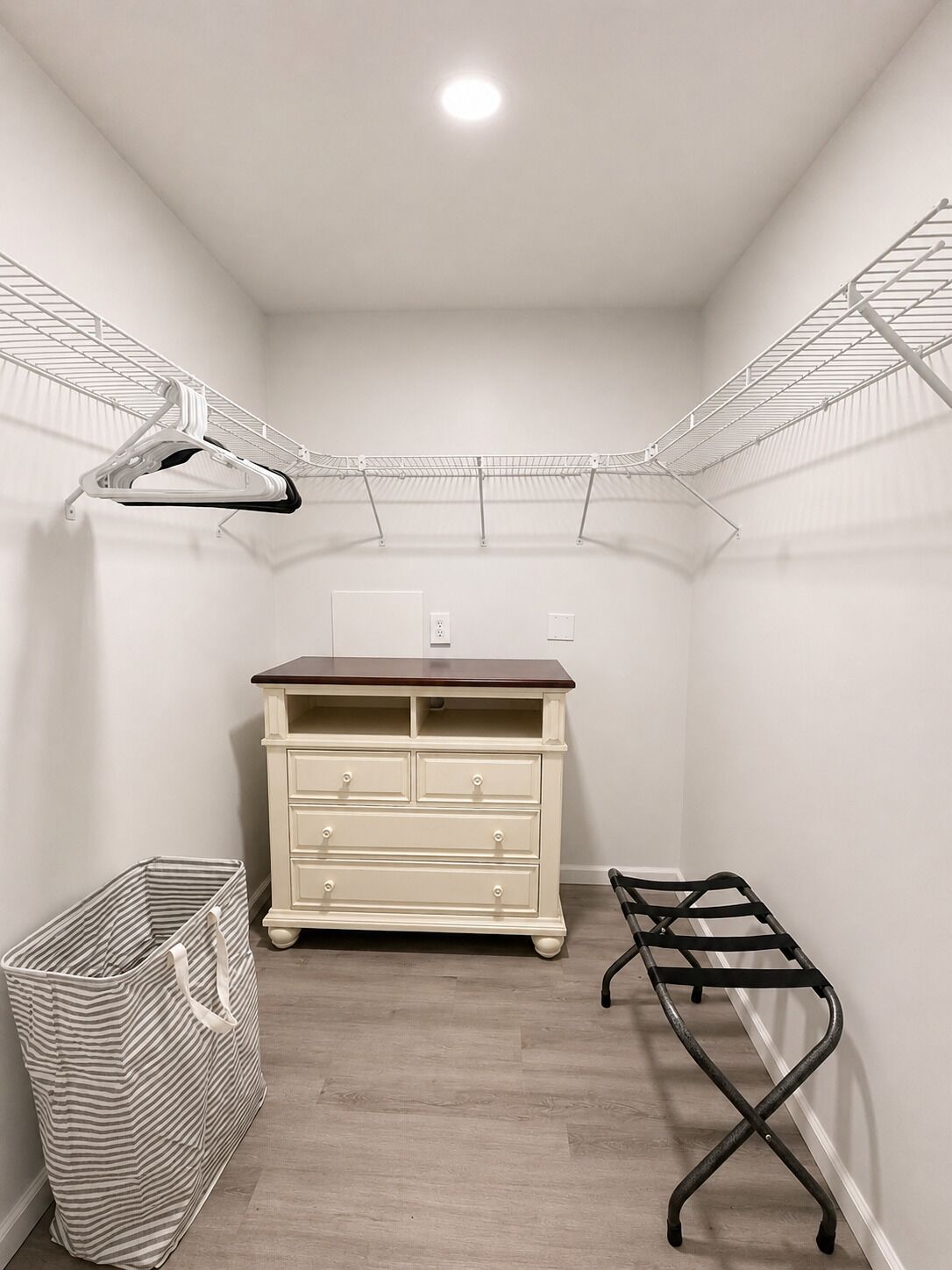 A corner closet with a wooden dresser, a metal drying rack, a striped laundry basket, and a metal shoe rack.