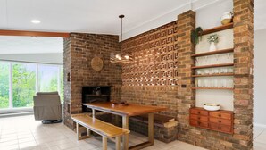 Un espace repas avec un mur en briques, une table en bois, un banc, un luminaire suspendu et des étagères où sont exposés des objets en verre.