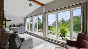 Un salon moderne doté d’une grande porte-fenêtre coulissante donnant sur un jardin, d’un canapé gris, d’un fauteuil en cuir marron et d’un coin cuisine avec un four à micro-ondes.