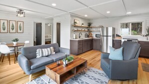 A modern living room with a grey sofa, a wooden coffee table, and a grey armchair. There is a kitchen area with dark cabinets and a stainless steel refrigerator. - Mountain View Cabin~HotTub~Fireplace~King Bed (New Market)