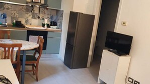 Eine kleine Küche mit einem Kühlschrank, einem Esstisch mit Stühlen und einem Fernseher auf einem Schrank.