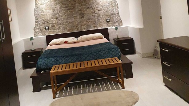 Ein Schlafzimmer mit einer Wand aus Stein, einem Bett mit blauer Bettdecke, Nachttischen, einer hölzernen Bank und einer Kommode.