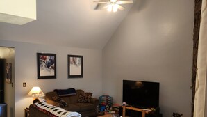 Una sala de estar con techo alto, un ventilador de techo, un televisor de pantalla plana, un sofá, una mesa de centro y una mesita de noche con lámpara.