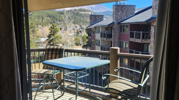 Un balcón con mesa y sillas, con vistas a un paisaje montañoso y edificios.