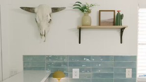 Une cuisine avec un plan de travail blanc, un bol de citrons, une lampe jaune, un crâne de vache, un cactus et un tableau encadré posé sur une étagère.