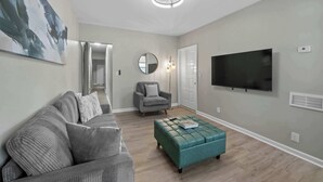 A modern living room with a grey sofa, teal ottoman, and a flat-screen TV mounted on the wall.