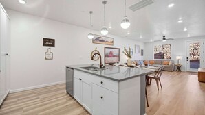 Una cocina moderna con una isla central, iluminación colgante y una zona de comedor con sofá y mesa.