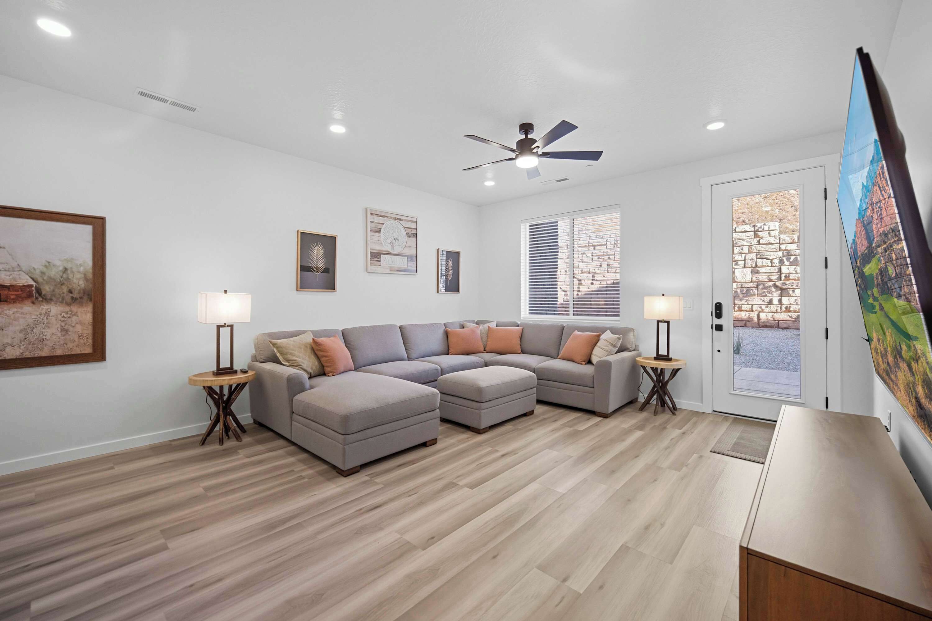 A modern living room with a sectional sofa, two side tables with lamps, a ceiling fan, and a large window with blinds.