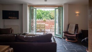 A living room with a brown leather sofa, a wooden coffee table, and a leather armchair. There is a television mounted on the wall, a fireplace, and a large window with a view of a garden and a brick wall.
