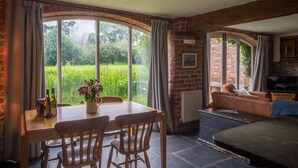 A cozy room with a dining table, chairs, a sofa, and a TV. Large windows offer a view of greenery outside.
