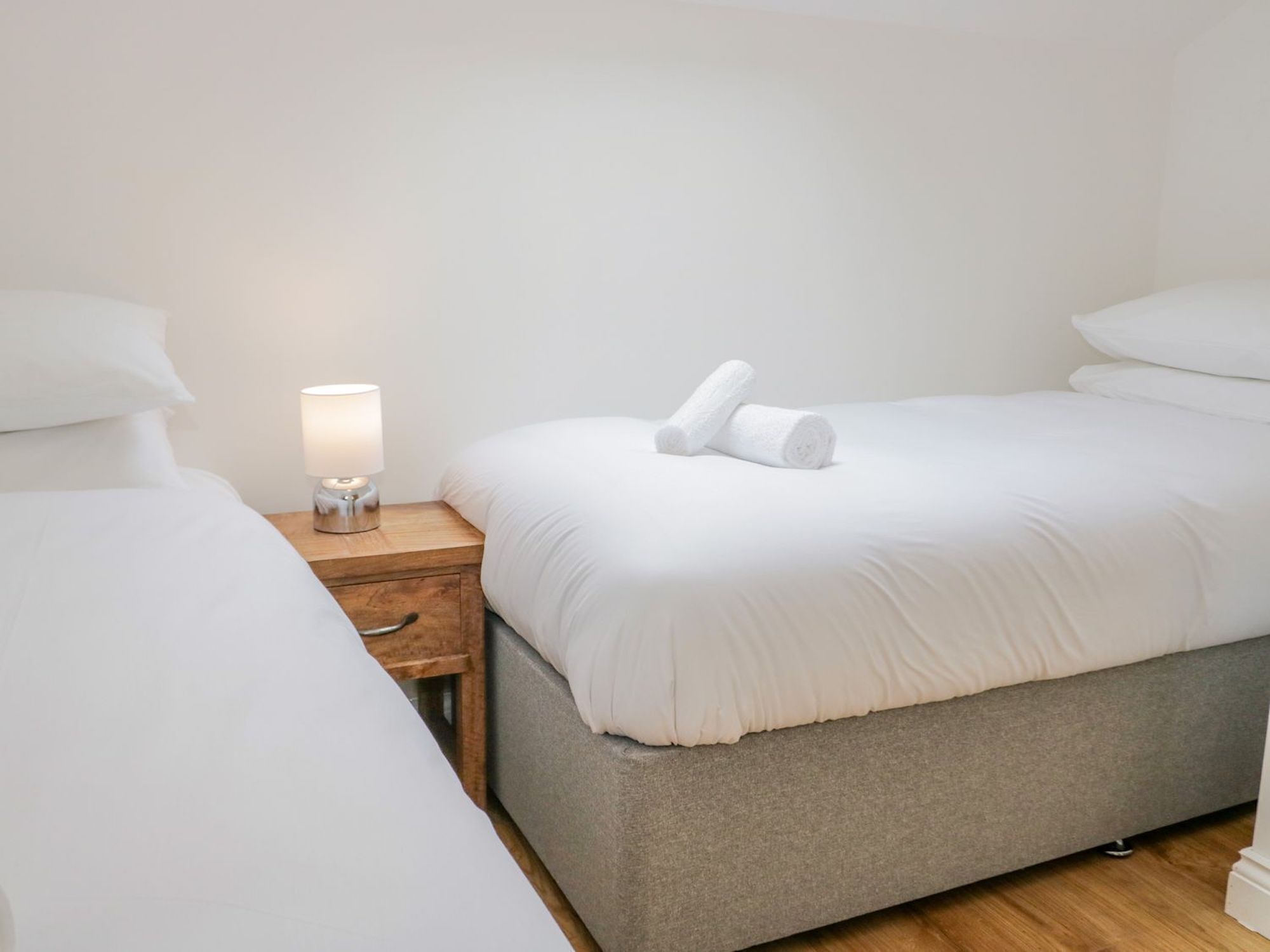 A hotel room with two single beds, a bedside table with a lamp, and a pair of rolled towels on the bed.