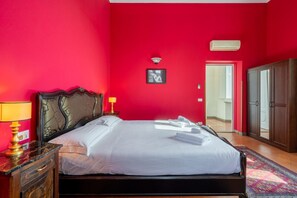 A hotel room with a red wall, a bed with white linens, a nightstand with a lamp, a wardrobe, and a door leading to another room. - Scale 50 Apartament (Firenze)