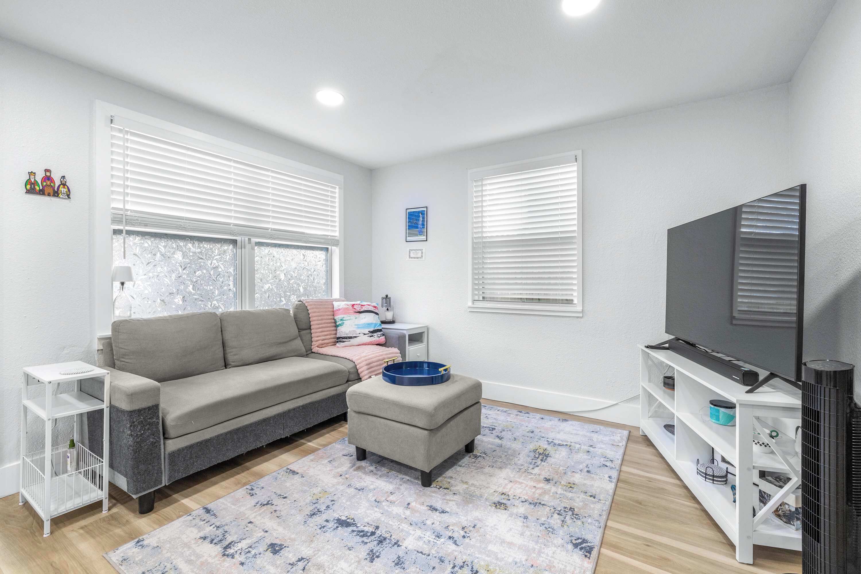 A modern living room with a grey sofa, a white side table, a blue ottoman, a TV stand, and a patterned rug.