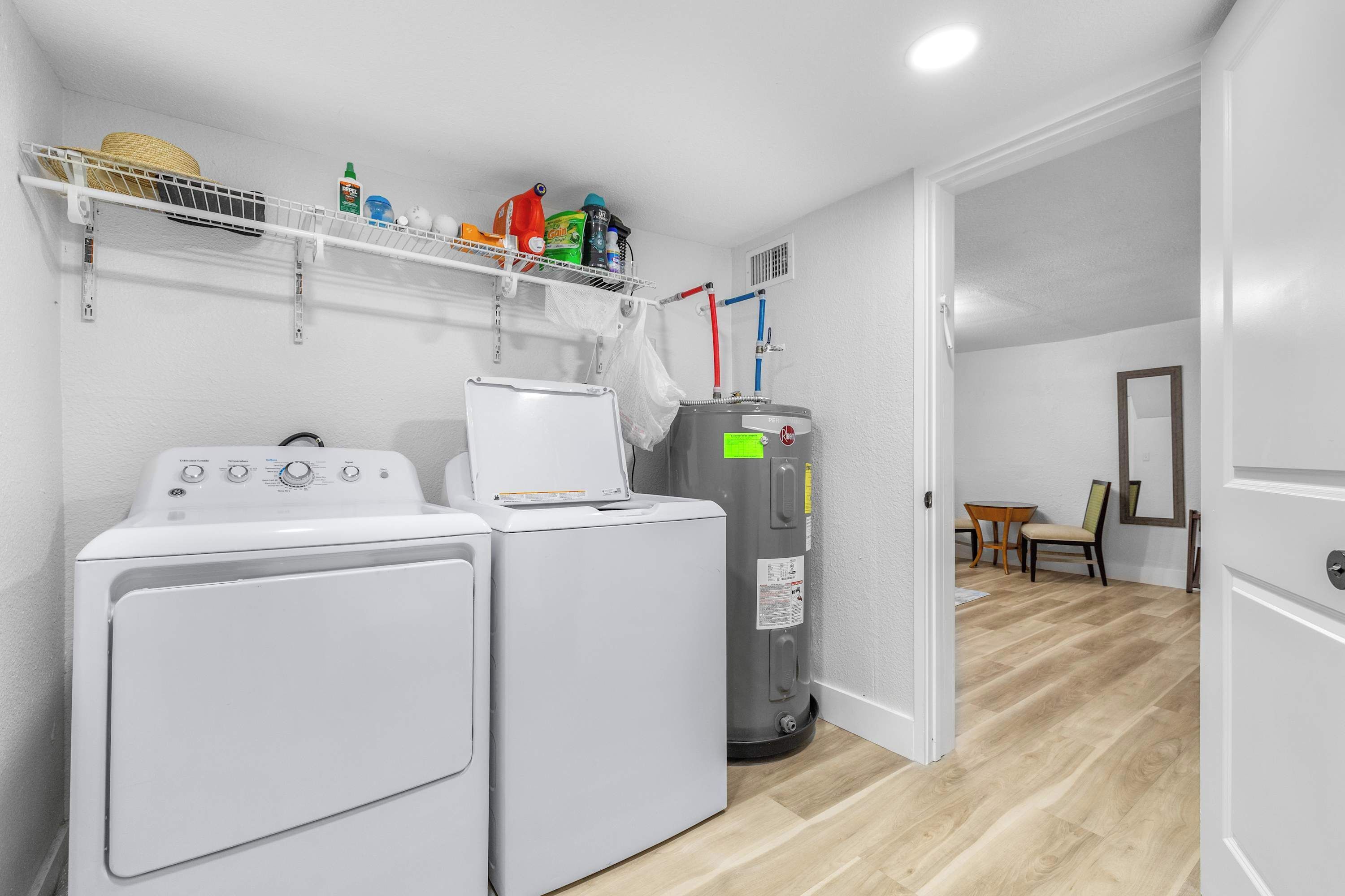 A laundry room with two washing machines, a dryer, and a water heater. There is a shelf above the machines holding various cleaning supplies.