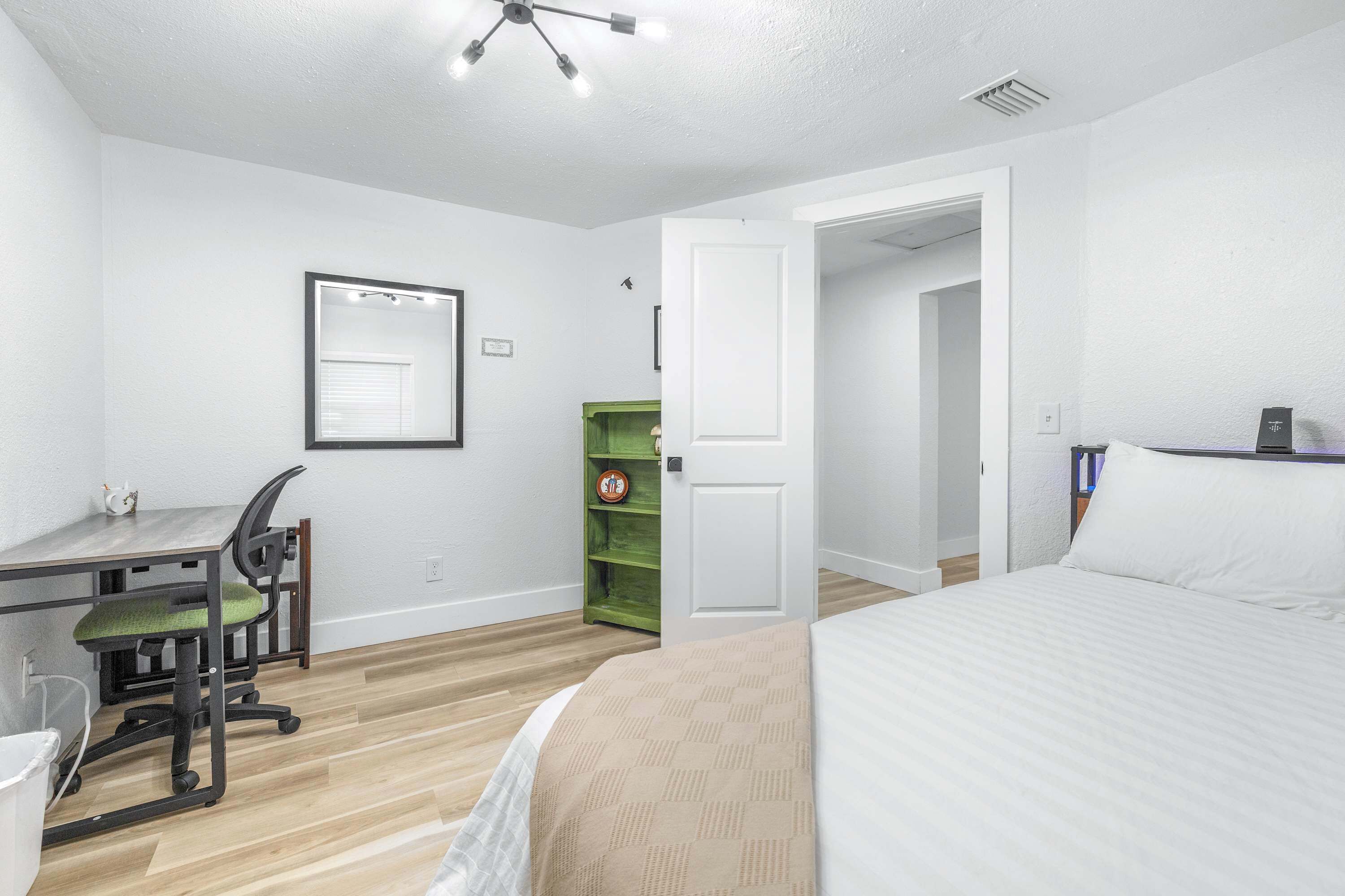 A bedroom with a bed, a desk with a chair, a green shelving unit, and a white door.