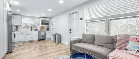 A modern apartment with a kitchen, a living area with a sofa and a coffee table, and a large window with blinds.