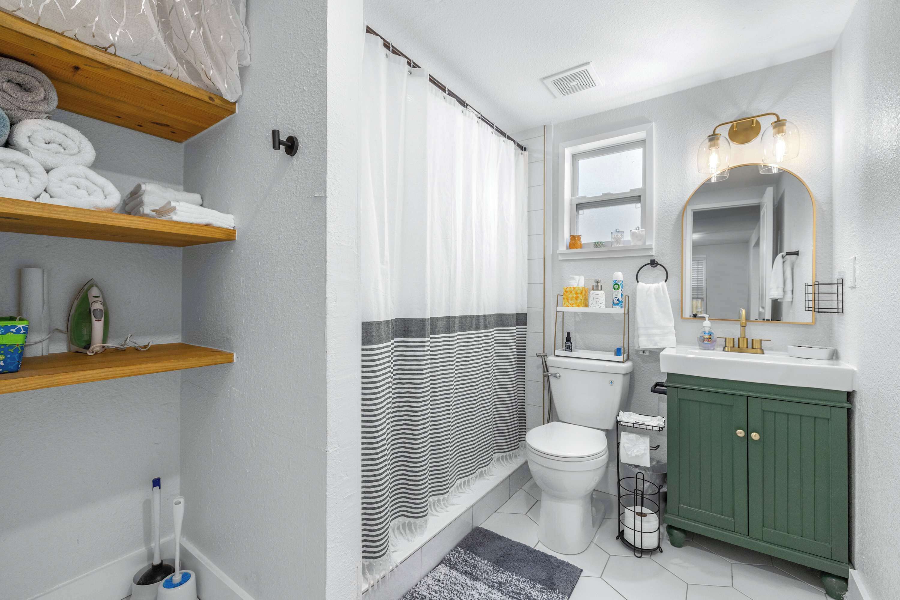 A bathroom with a shower, toilet, and green vanity. There are shelves with towels and toiletries.