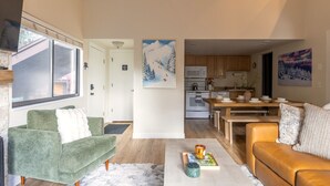 Uma sala de estar moderna com um sofá verde, um sofá de couro marrom e uma mesa de centro. Há uma área de cozinha com armários de madeira e uma mesa de jantar.