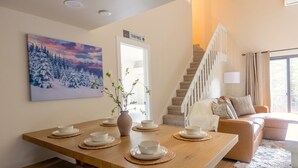 Uma sala de jantar com uma mesa de madeira posta para quatro pessoas, uma escada e uma sala de estar com um sofá.