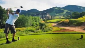 Um campo de golfe com um jogador em pleno movimento, céu limpo e paisagem acidentada ao fundo.