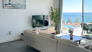 Un soggiorno moderno con una grande TV, un divano, un tavolino e vista mare attraverso ampie finestre.