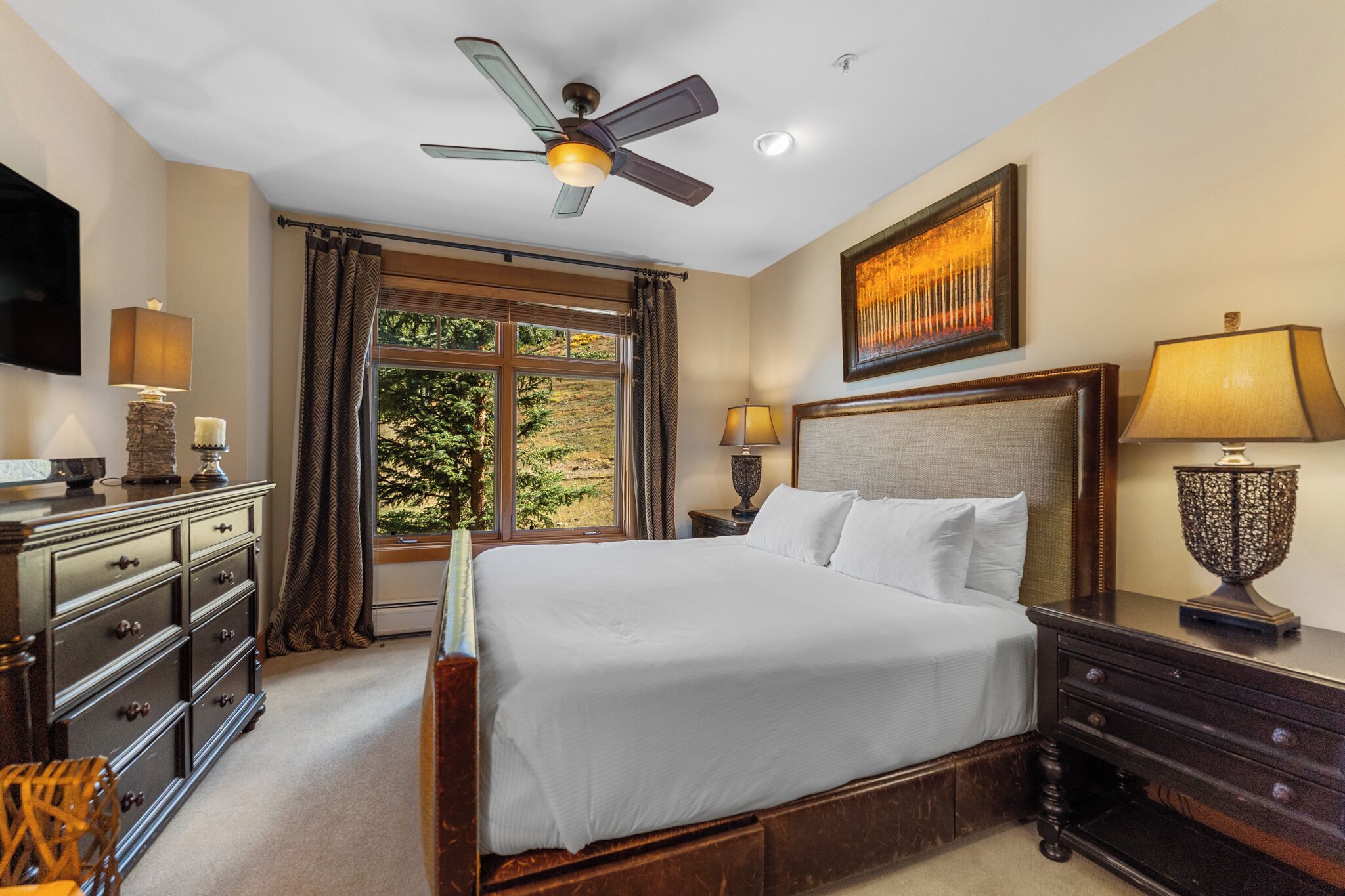 A well-lit bedroom with a large bed, a ceiling fan, a TV, and a window with curtains.