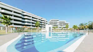 A swimming pool with a tiled deck, surrounded by a fence, and adjacent to a modern multi-story building with balconies.