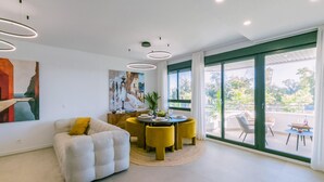 A modern living room with a sofa, dining table, and chairs, large windows with a view, and artwork on the walls.