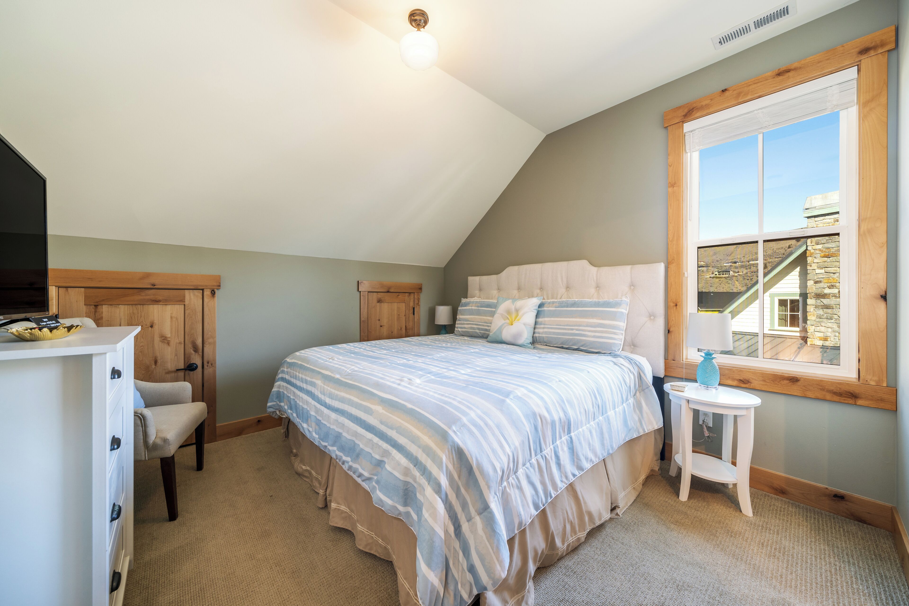 Une chambre avec un plafond en pente, un lit recouvert d’une couvre-lit à rayures, une table de chevet avec une lampe et une fenêtre donnant sur une maison.