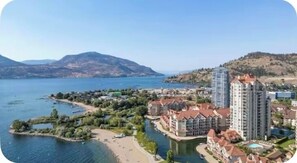 Eine Küstenstadt mit einer weiten Wasserfläche, einem Fluss und einem Berg im Hintergrund. Es gibt Wohngebäude und ein hohes Hotel mit rotem Dach.