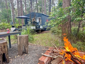 Una cabaña en un bosque con un hoyo para la fogata y mobiliario exterior.