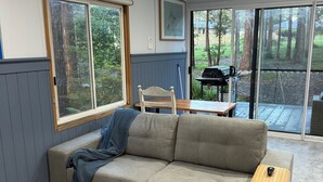 Un salon moderne avec un canapé gris, une table basse en bois et une table à manger avec des chaises. De grandes fenêtres offrent une vue sur l’extérieur.