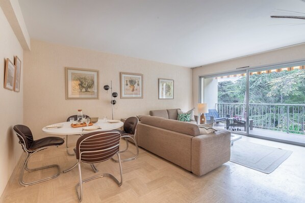 Un salon moderne comprenant un canapé, deux fauteuils, une table à manger avec un plateau en verre et un balcon offrant une vue sur la verdure.