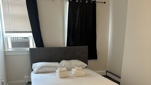 A neatly made bed with white linens and two towels, a grey upholstered headboard, and a window with blinds and a black curtain. - Stylish Apt: 20 Min to Manhattan, Groups (West New York)