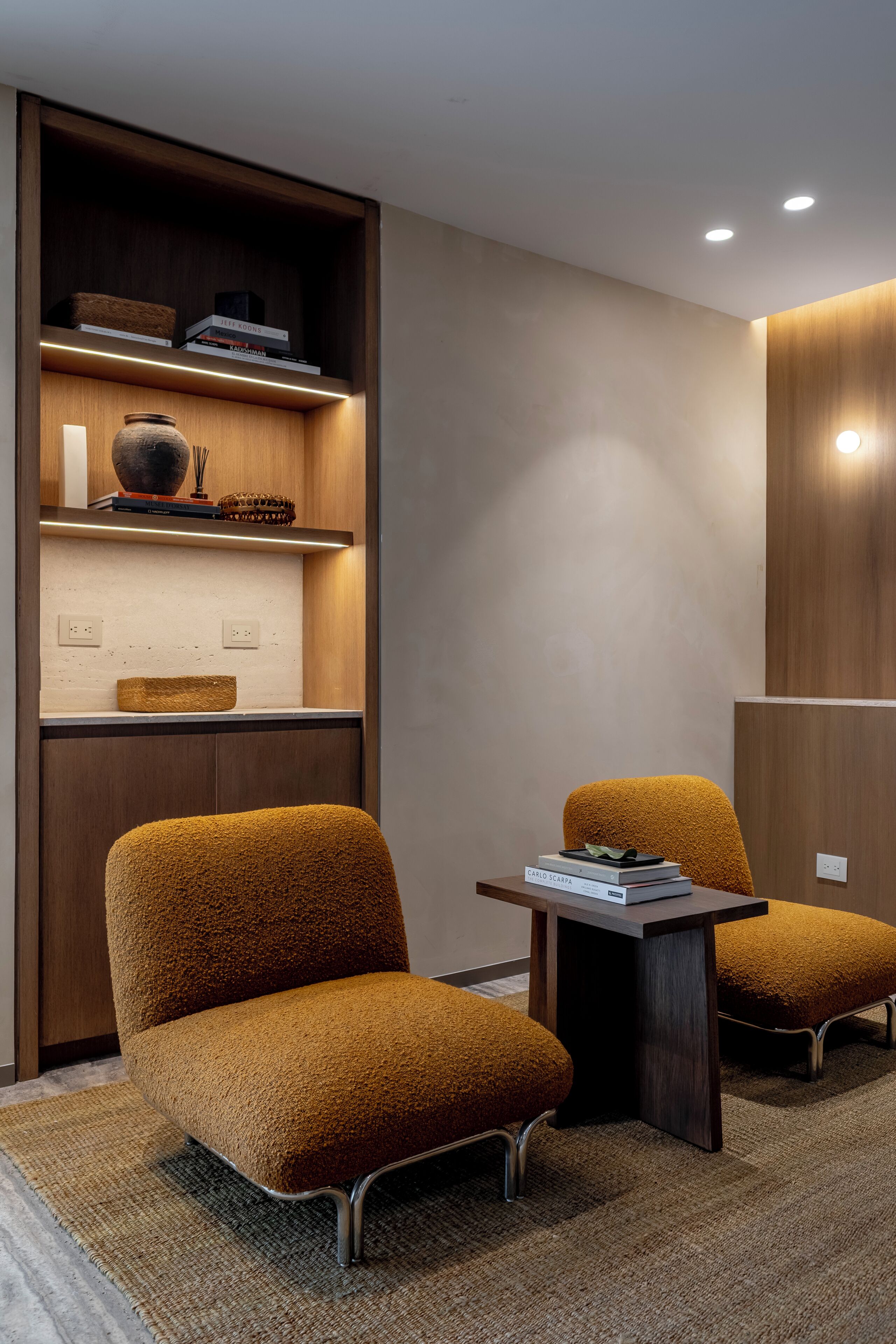 Une pièce moderne avec deux fauteuils marron à revêtement en tissu, une petite table et une étagère en bois où sont disposés des livres et des objets décoratifs.