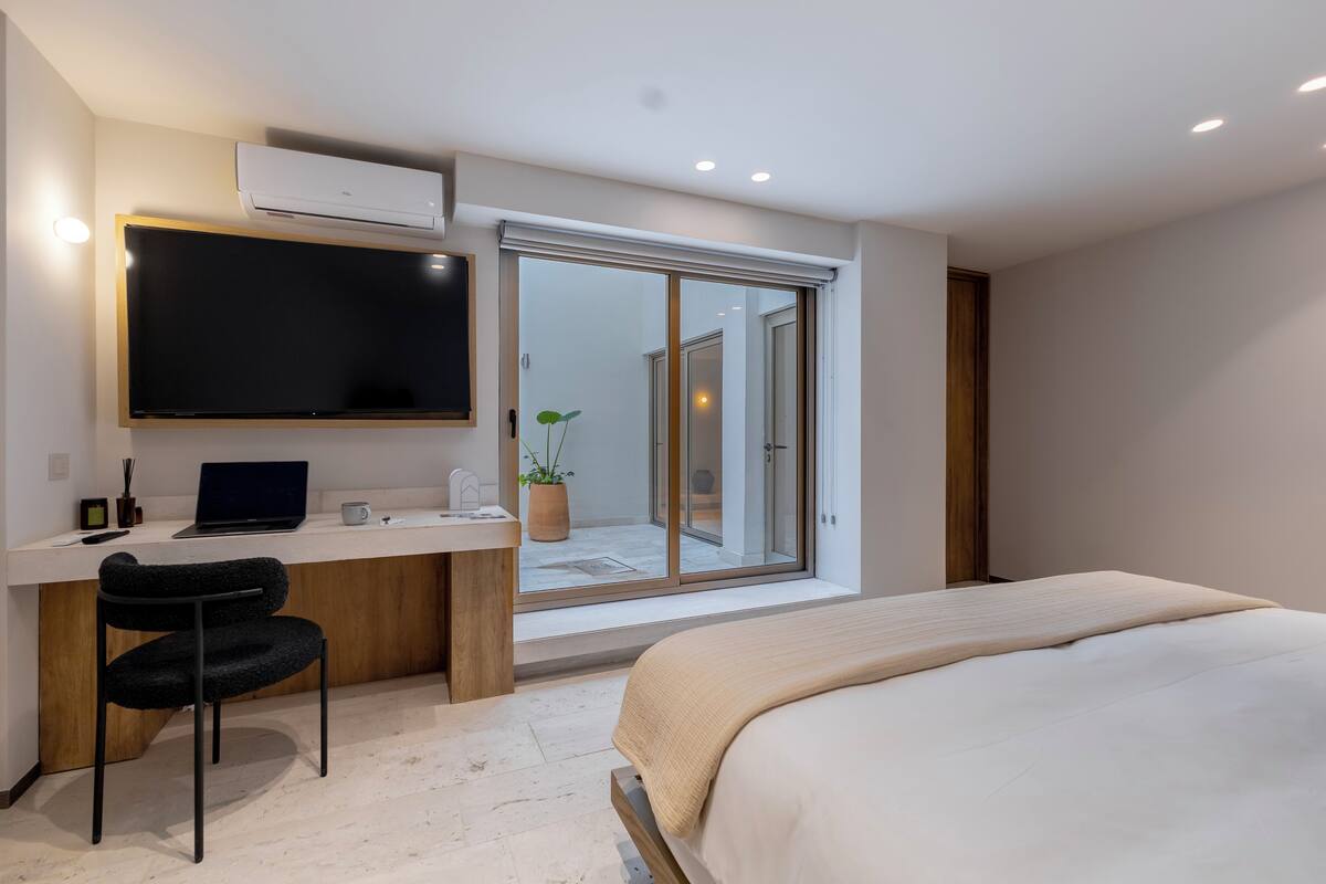 A modern bedroom with a bed, a desk with a laptop, a chair, and a sliding glass door leading to a balcony.