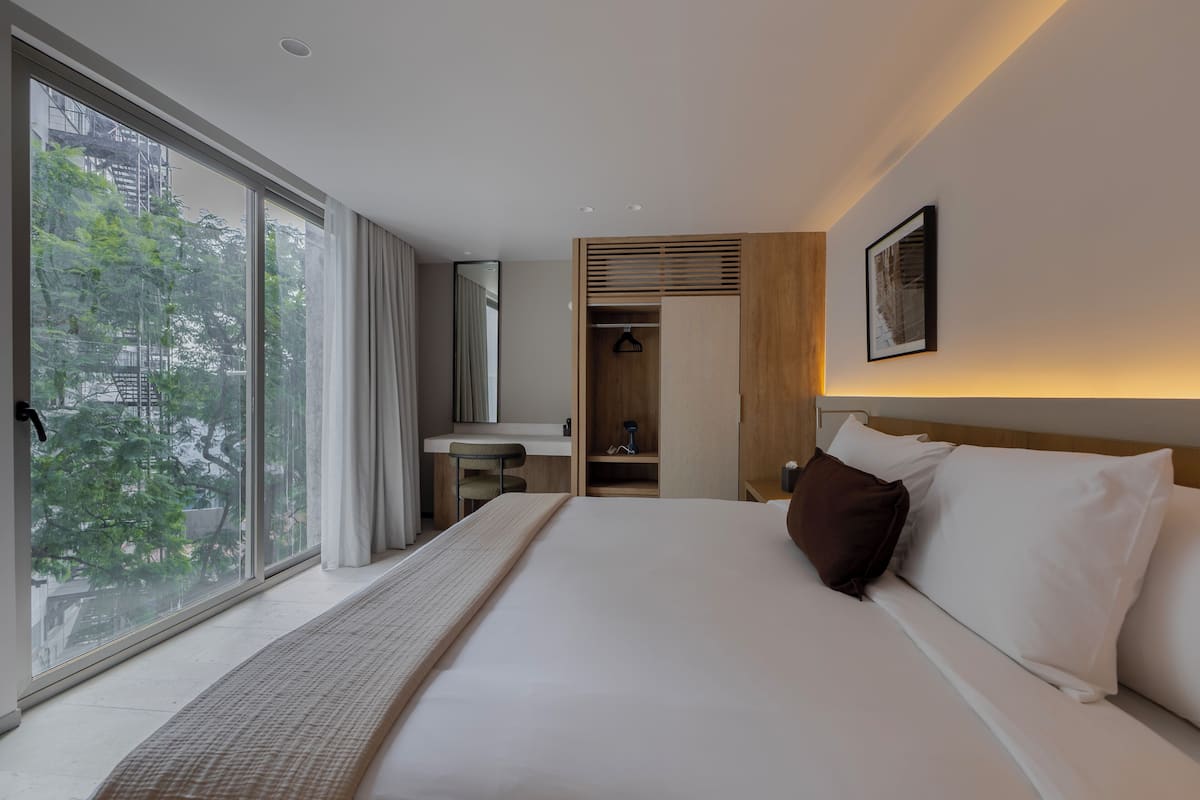 A hotel room with a large window, a bed with white linens, a wooden wardrobe, and a small desk with a mirror.